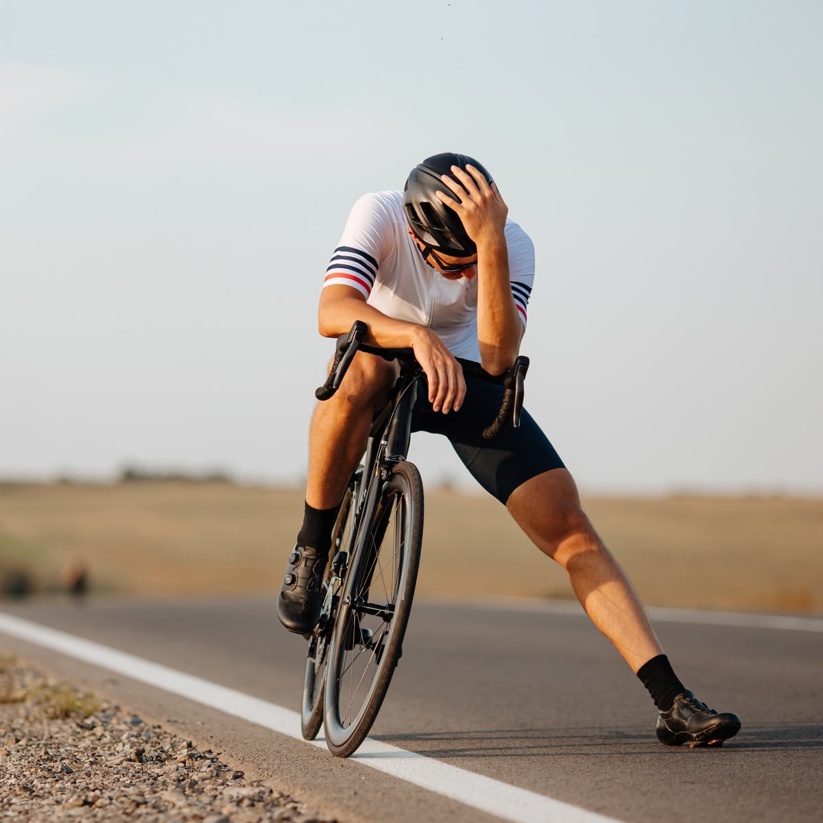 A cyclist wearing a helmet and sport clothes sits slumped on his bike at the roadside, head in hand, looking exhausted or distressed on an empty rural road.