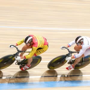 Two cyclists racing on a velodrome track, one in a yellow and red suit and the other in white with blue and red details. The image captures their speed with motion blur.