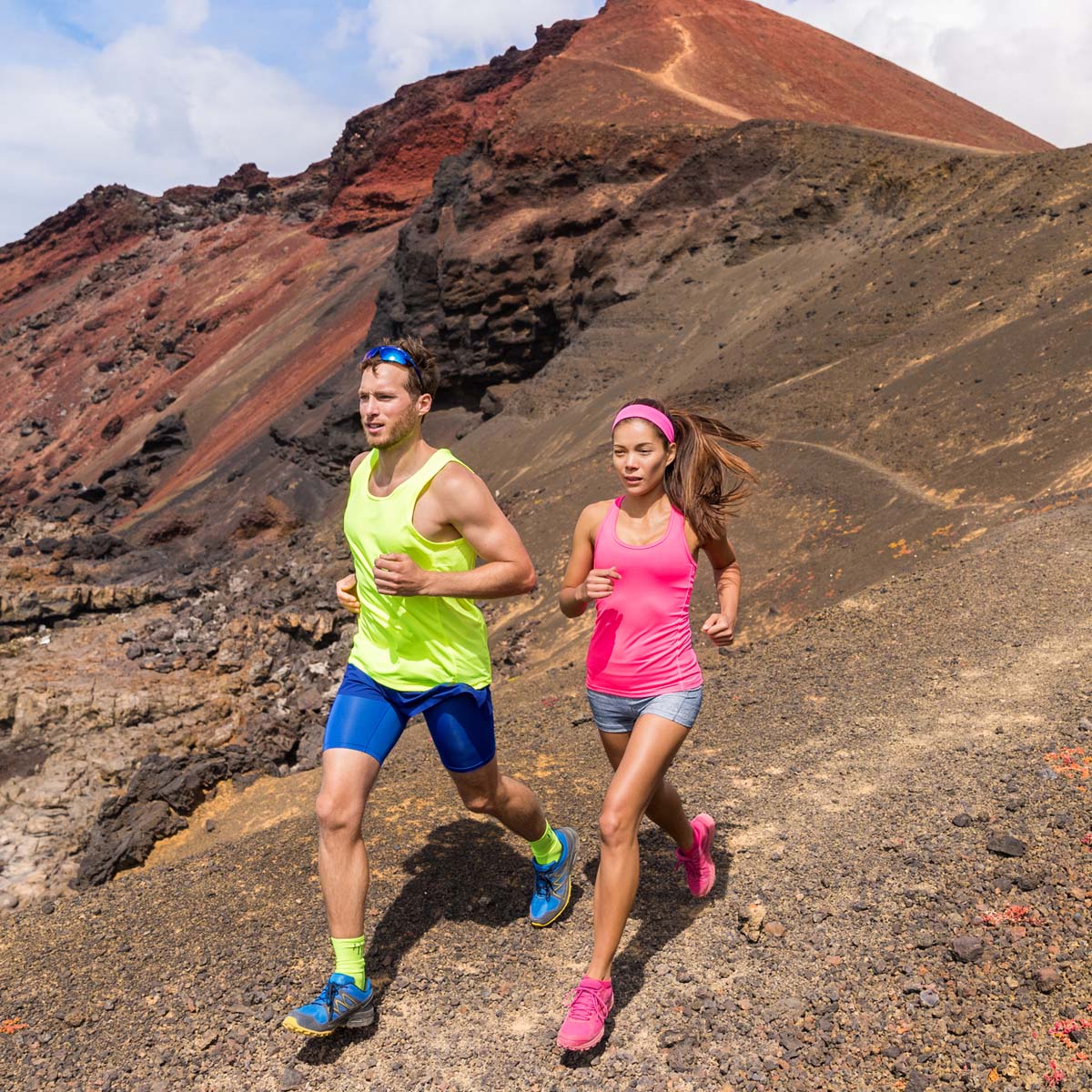 A man and a woman in bright athletic wear run on a rocky, volcanic trail with red and black hills behind them, showcasing the adventure of an endurance training camp.