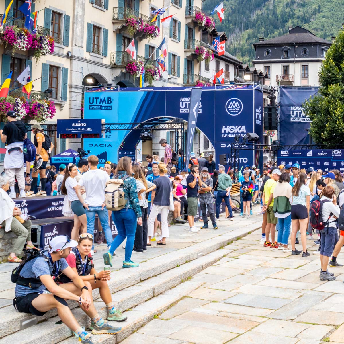 A large crowd gathers at the UTMB ultra running event in a French mountain town, with banners and flags displayed, buildings decorated with flowers, and forested mountains in the background.