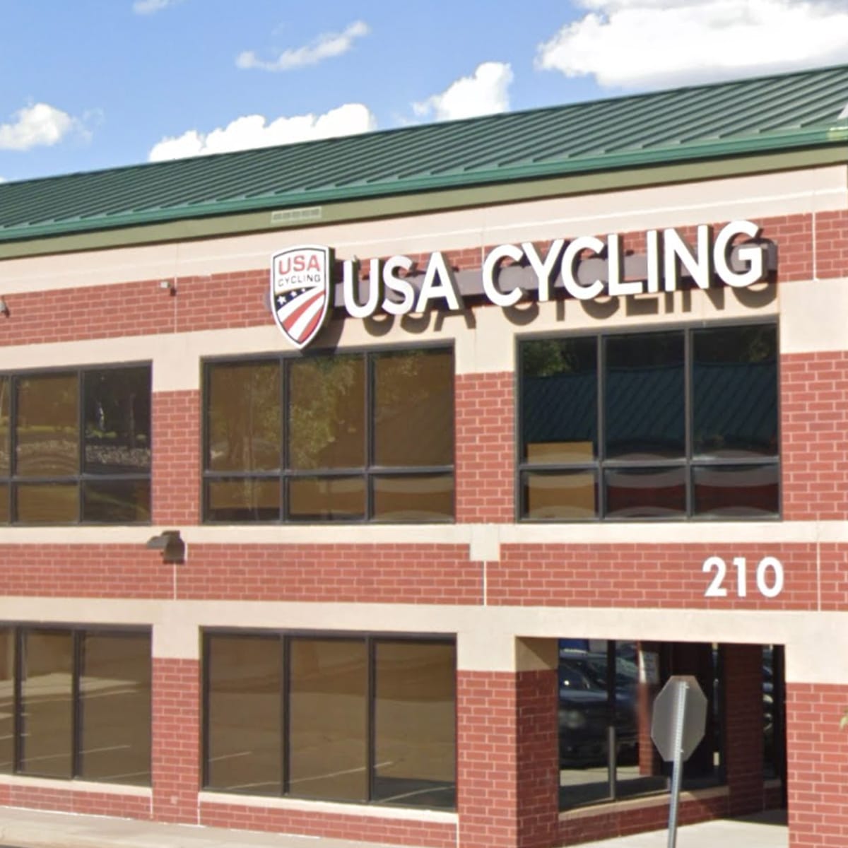 A brick building with USA Cycling signage and logo is shown, address number 210 near the entrance.