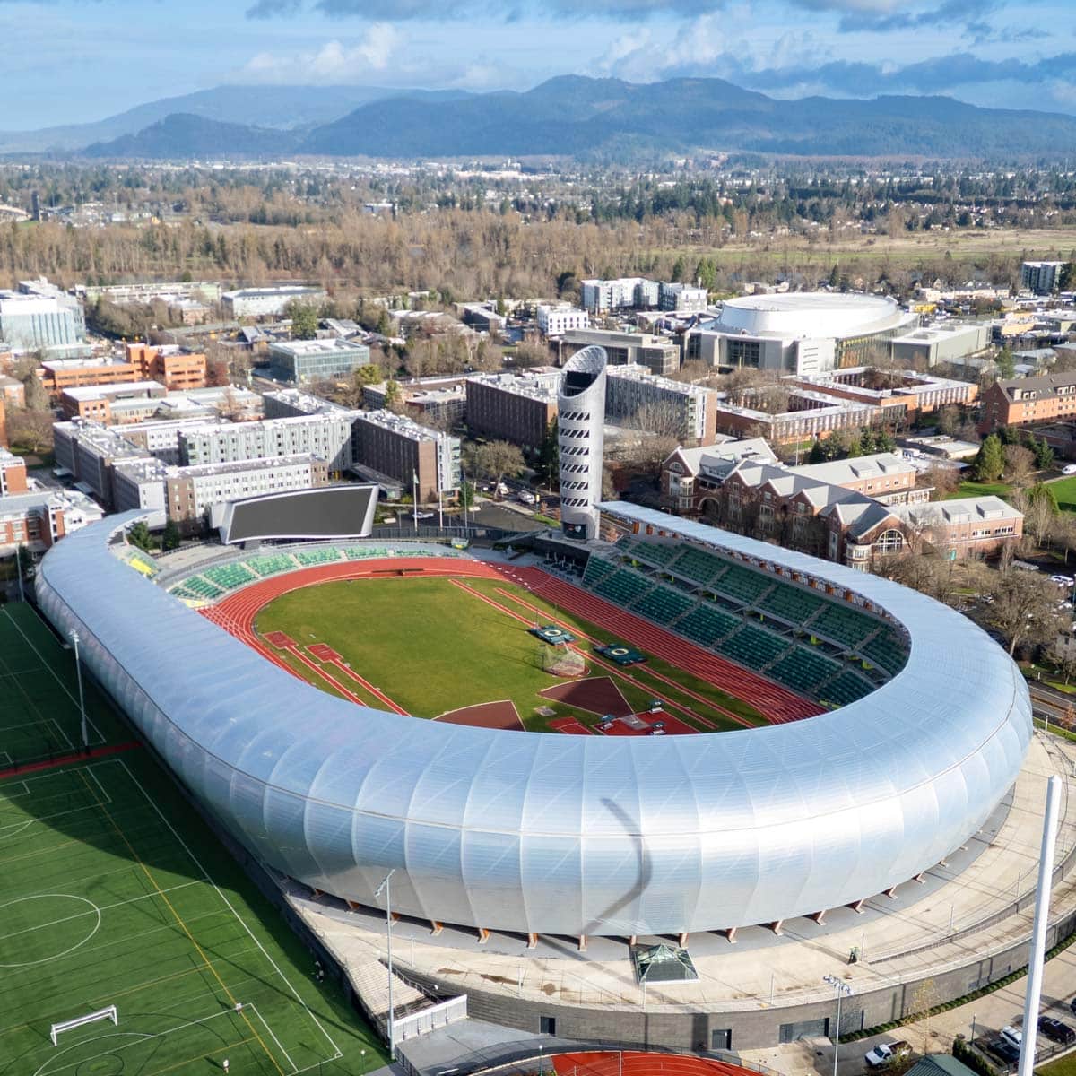 Aerial view of the Hayward Field outdoor stadium dedicated to track and field, surrounded by city buildings and trees, with mountains in the background under a partly cloudy sky—a stunning setting for USA athletics events.