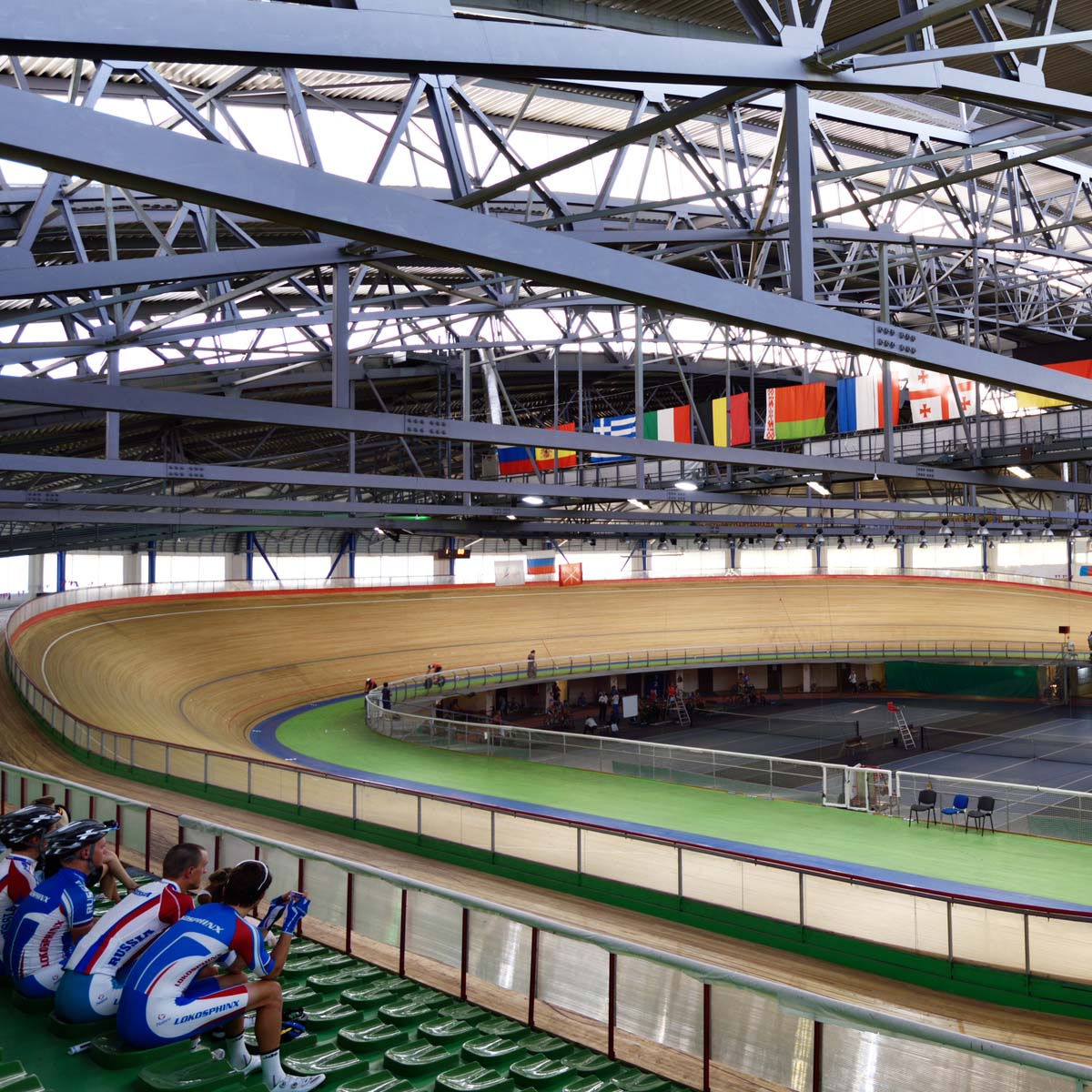 An indoor velodrome with a wooden cycling track, steel beams overhead, and international flags. Spectators sit in the foreground.