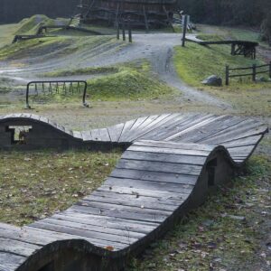 A wooden mountain bike washboard feature with undulating, wave-like sections stands in a grassy outdoor park with gravel paths, trees, and other bike structures.