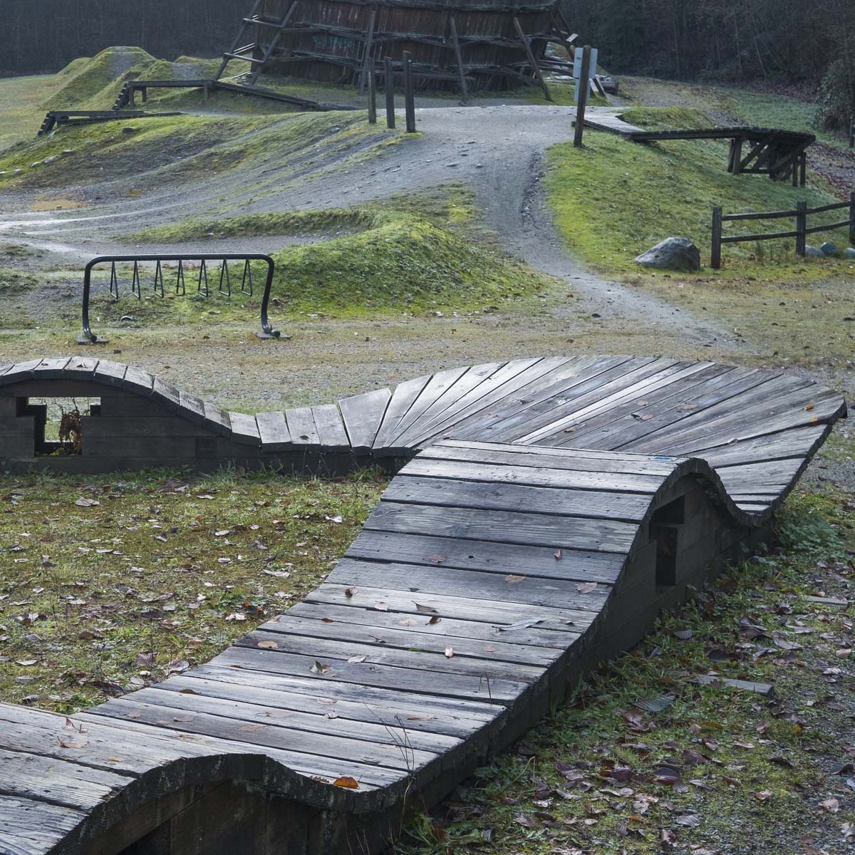A wooden mountain bike washboard feature with undulating, wave-like sections stands in a grassy outdoor park with gravel paths, trees, and other bike structures.
