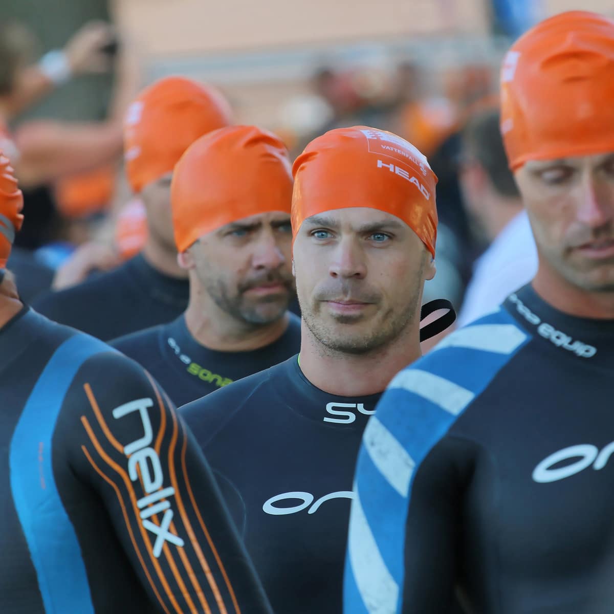 A group of athletes in orange swim caps and triathlon wetsuits stand in line, preparing for a Wetsuit Legal swimming event. The focus is on a man in the center, with others slightly out of focus around him.