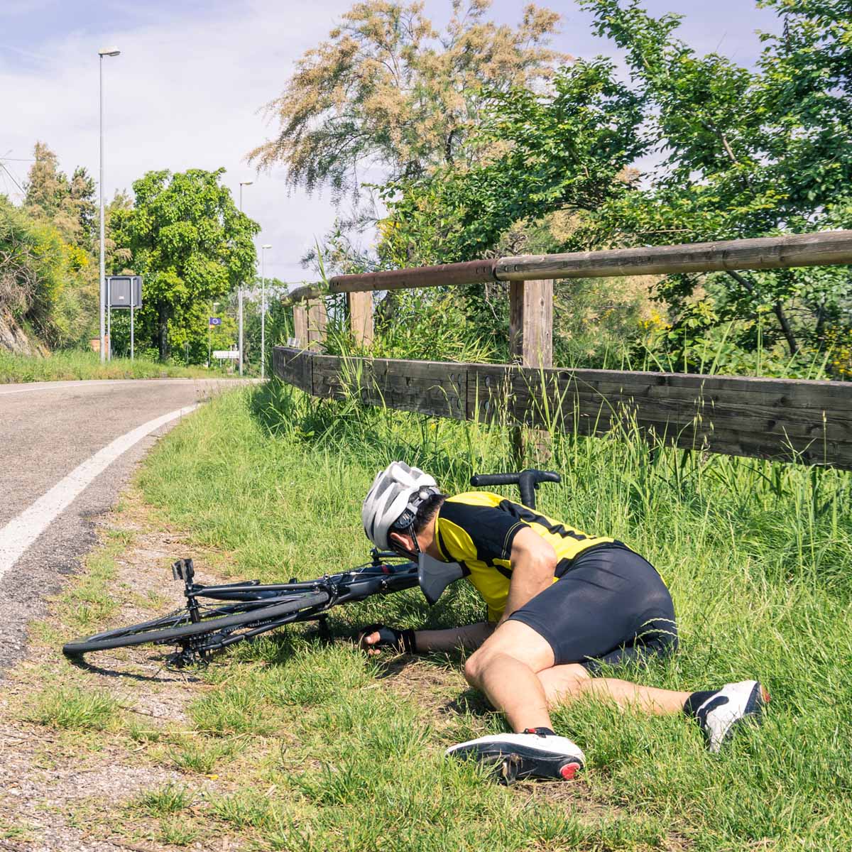 A cyclist in a yellow and black outfit has fallen off his bike onto the grassy roadside, lying face down with his helmet on. His bicycle rests on its side next to him near a wooden fence.