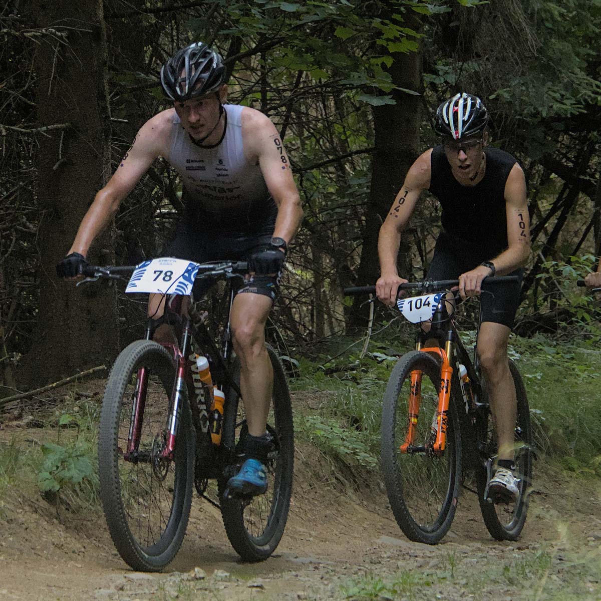Two male cyclists wearing race numbers ride mountain bikes along a forest trail during an XTERRA competition. They are focused and riding closely together, surrounded by trees and greenery.