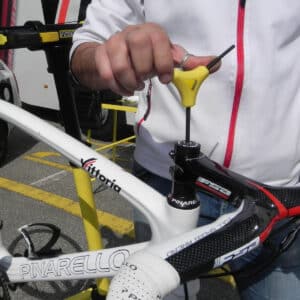 A person uses a yellow Y-shaped wrench to adjust the stem of a white Pinarello road bike, wearing a white jacket with red trim. This moment unfolds outdoors on a sunny day during a cycling race.