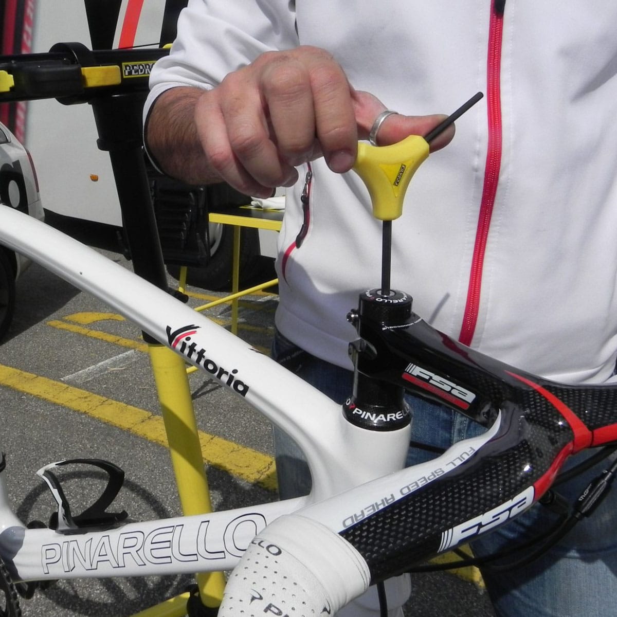 A person uses a yellow Y-shaped wrench to adjust the stem of a white Pinarello road bike, wearing a white jacket with red trim. This moment unfolds outdoors on a sunny day during a cycling race.