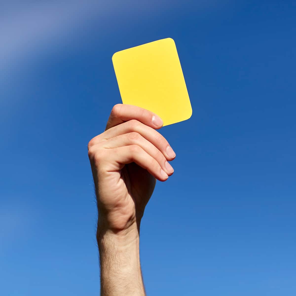 A hand holds up a yellow card against a clear blue sky, suggesting a warning or caution, often seen in sports events and competitions.