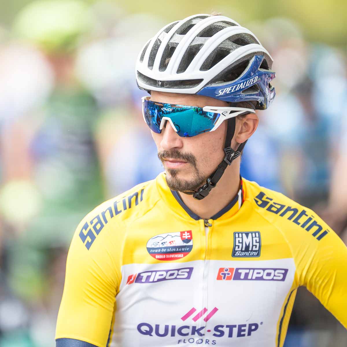 A male cyclist wearing a yellow Santini jersey with sponsor logos, helmet, and sunglasses stands outdoors.