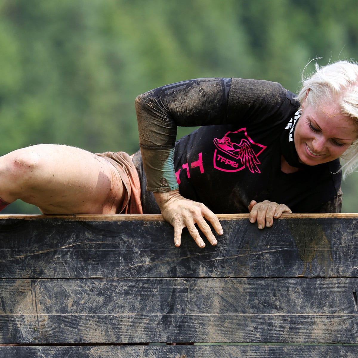 A woman with mud on her arms and legs climbs over a wooden wall during a Spartan outdoor obstacle course, smiling as she exerts effort. Green, blurry trees are visible in the background.
