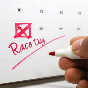 A calendar page with the 25th date boxed and marked in red. Race Day is written in large script below. A hand with a red marker writes on the calendar.