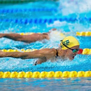 A competitive swimmer in a yellow cap with a Swedish flag performs the butterfly stroke in a pool, with another swimmer behind. Blue and yellow lane markers are visible.