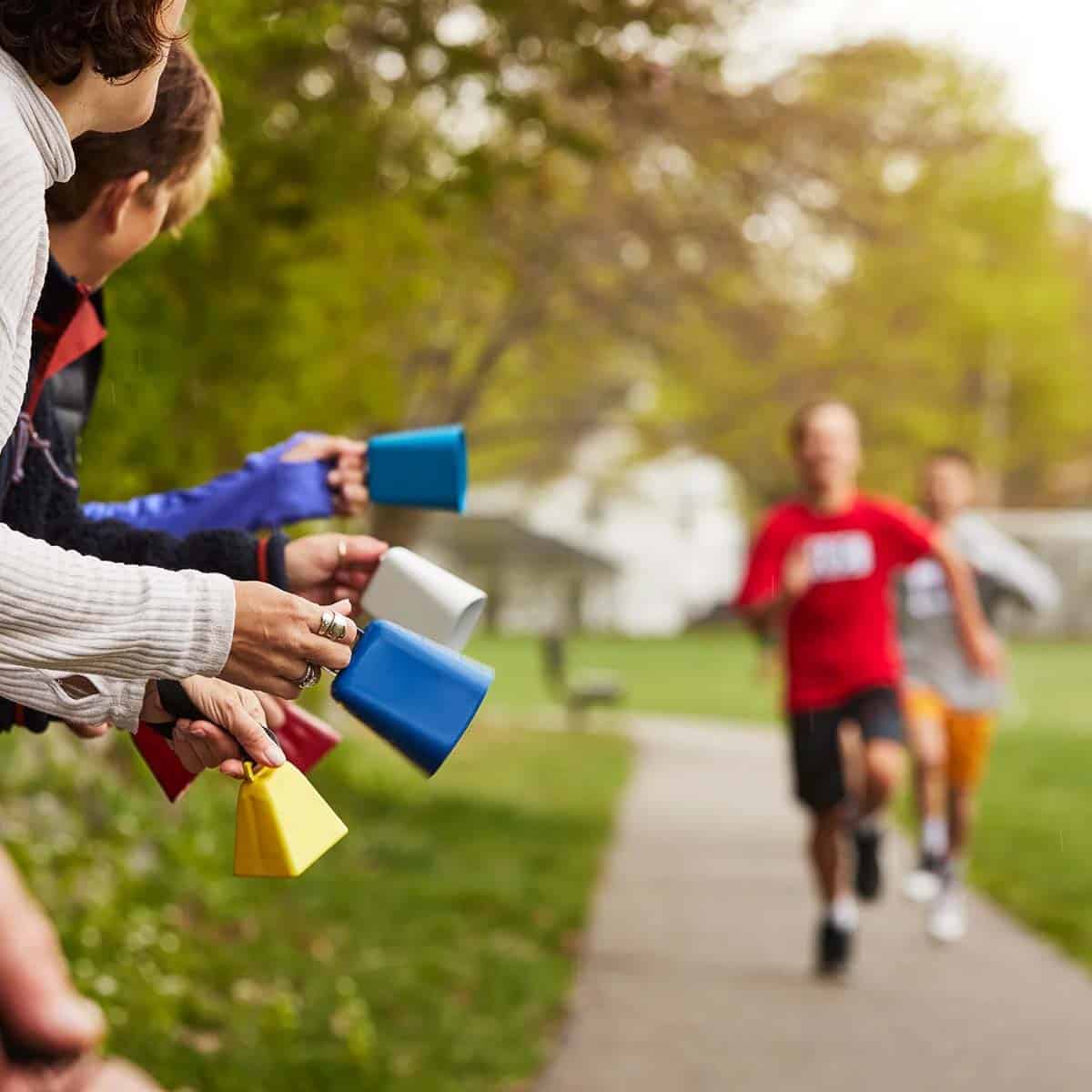 People ring colorful cowbells while cheering on runners during a race along a tree-lined path, capturing the supportive spirit celebrated in outdoor sporting events.