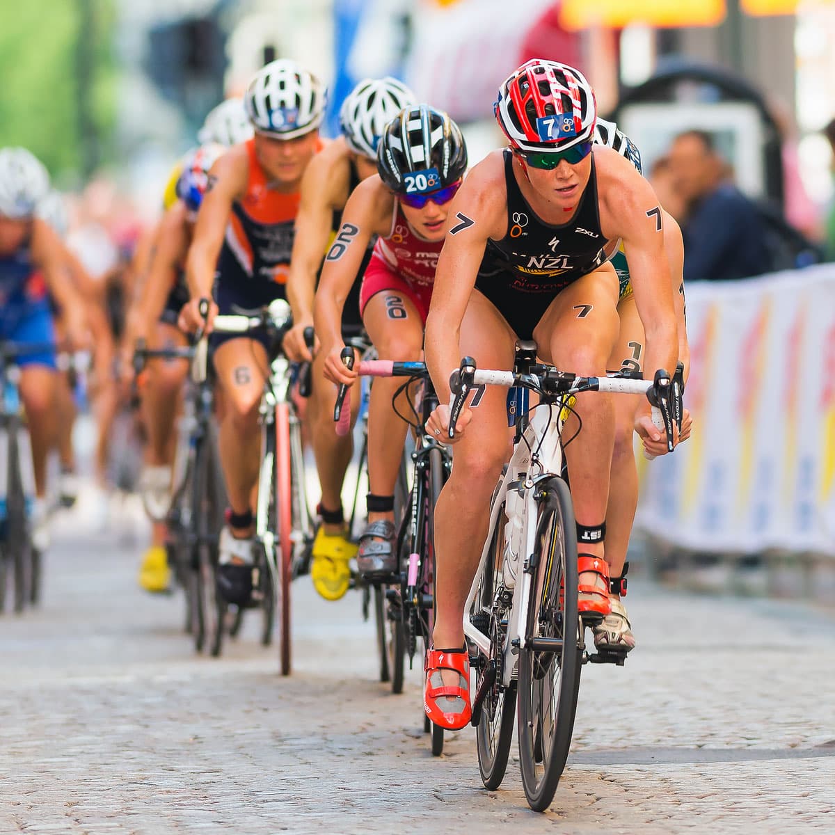 A group of focused triathletes wearing helmets and race numbers compete closely on bicycles during a race, with a city street and blurred spectators in the background.