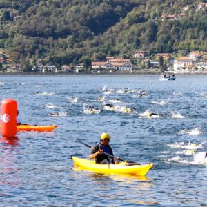 Athletes swim in open water near a lakeside town as kayakers escort them; hills with houses rise in the background. A red buoy marks the race course.