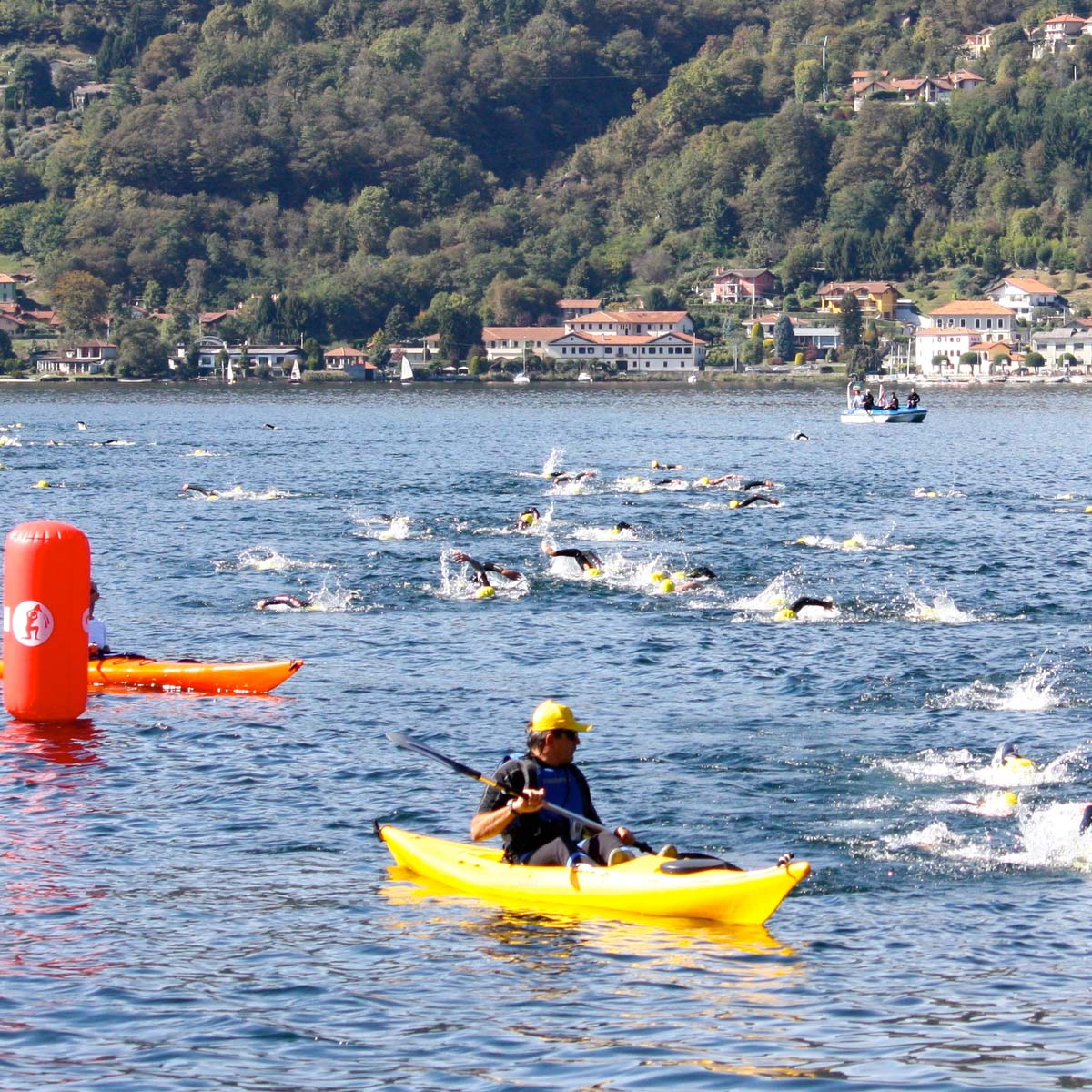 Athletes swim in open water near a lakeside town as kayakers escort them; hills with houses rise in the background. A red buoy marks the race course.