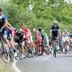 Cyclists from various teams take a nature break by the roadside during a race, with team support cars, spectators, and bikes visible among trees and greenery.
