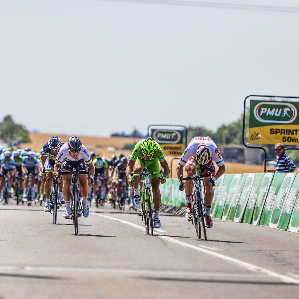 Cyclists race at high speed toward the finish line, with two leading riders neck and neck. Signs read Sprint 50m, and a group of racers follows close behind under the sunny sky—an exciting moment featured on Enduropedia.