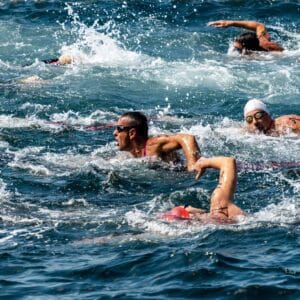 Several swimmers wearing goggles and swim caps compete in open water, surrounded by splashing waves.