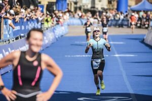 Jacqueline Busse, Ho'Omau Director of Global Operations, smiles and gestures while running toward the finish line on a blue track at her first Olympic distance triathlon in Hamburg, Germany, with other athletes and cheering spectators in the background.