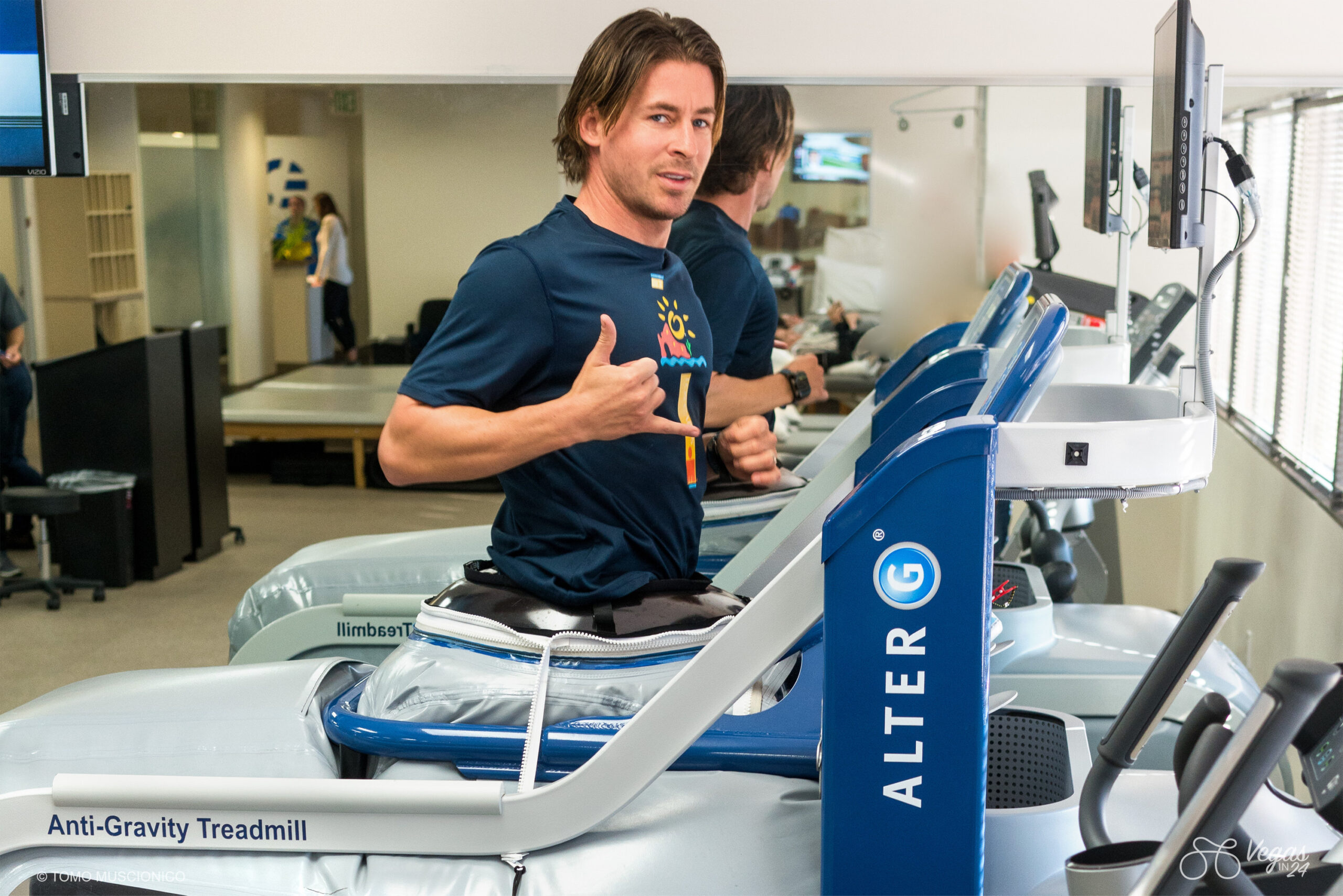 Andy Funk, Chief of Ho'Omau Endurance Co, exercises on an AlterG Anti-Gravity Treadmill in a gym, giving a double thumbs-up and looking toward the camera, proudly training for his epic 365-mile long Vegas In 24 ultra-cycling ride, with mirrors and gym equipment in the background.