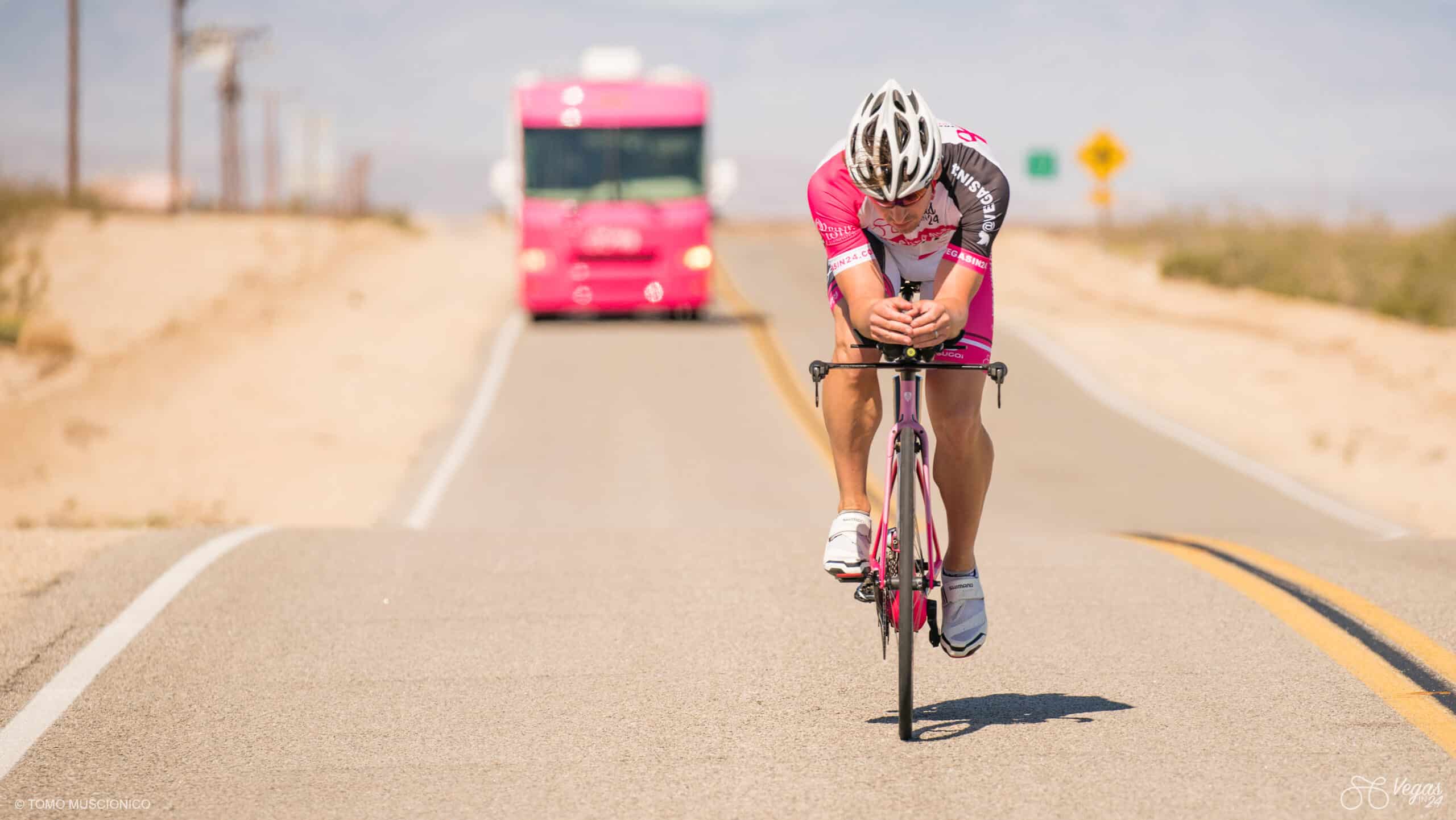 Andy Funk, Chief of Ho'Omau Endurance Co., in pink racing gear and helmet rides on an empty desert road during the original Vegas In 24 ultra-cycling challenge, closely followed by a pink RV, under a clear sky.