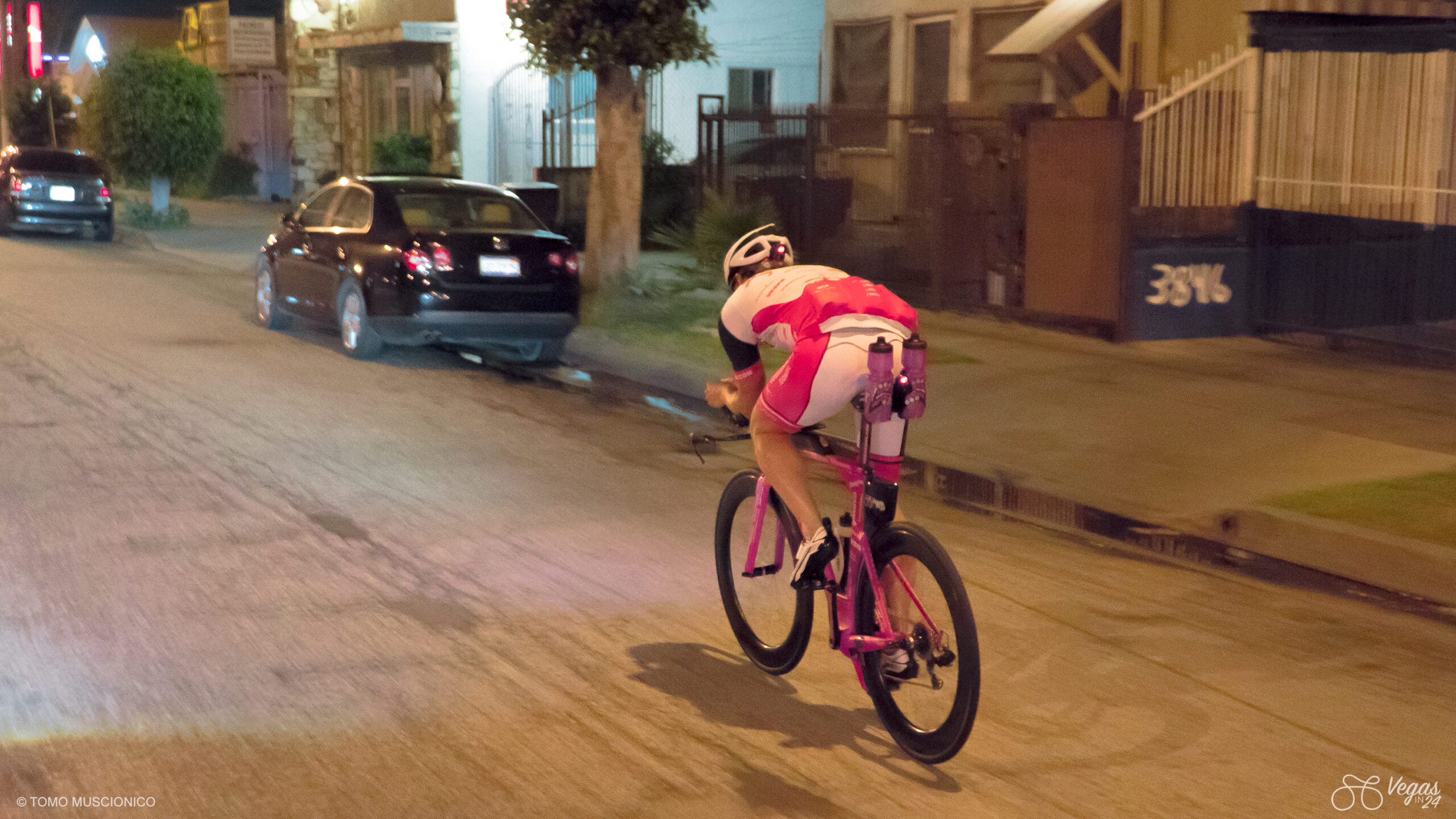 Andy Funk, Chief of Ho'Omau Endurance and creator of the original Vegas In 24 ultra-cycling challenge, rides a pink bike in a pink and white outfit down a dimly lit urban street in Campton at night, passing parked cars and houses with barred windows.