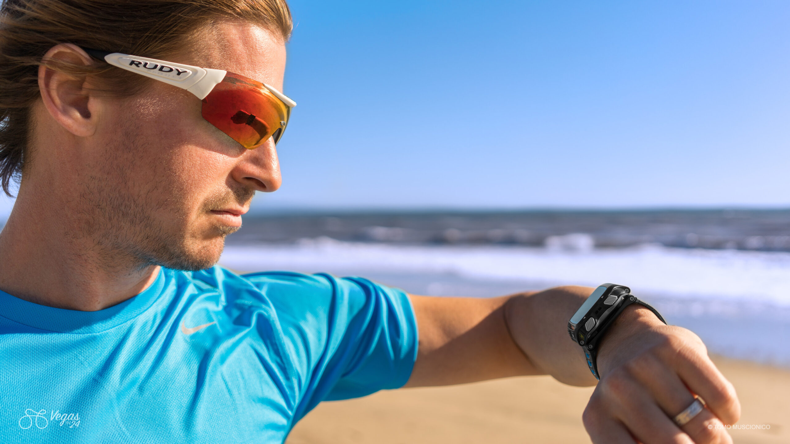 Andy Funk, Chief of Ho'Omau Endurance, wearing Rudy Project sport sunglasses and a blue running shirt checks his Garmin smartwatch on a sunny beach, ocean in the background, during one of his last training runs prior to the original Vegas In 24 ultra-cycling challenge.