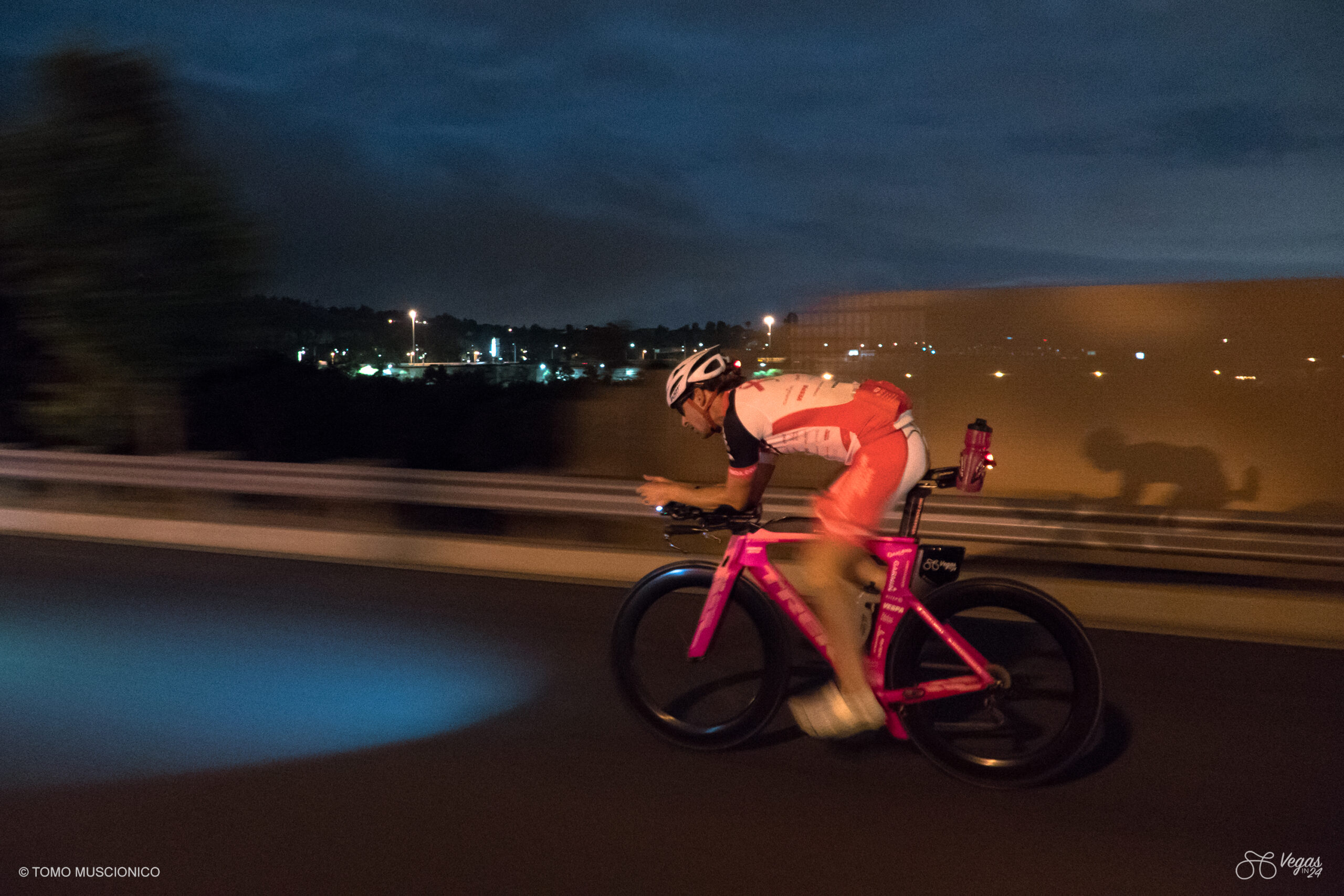 Andy Funk, Chief of Ho'Omau Endurance, in a pink racing outfit and helmet speeds down the road in the dark during the early hours of the original Vegas In 24 ultra-cycling challenge, city lights shining beneath the night sky.