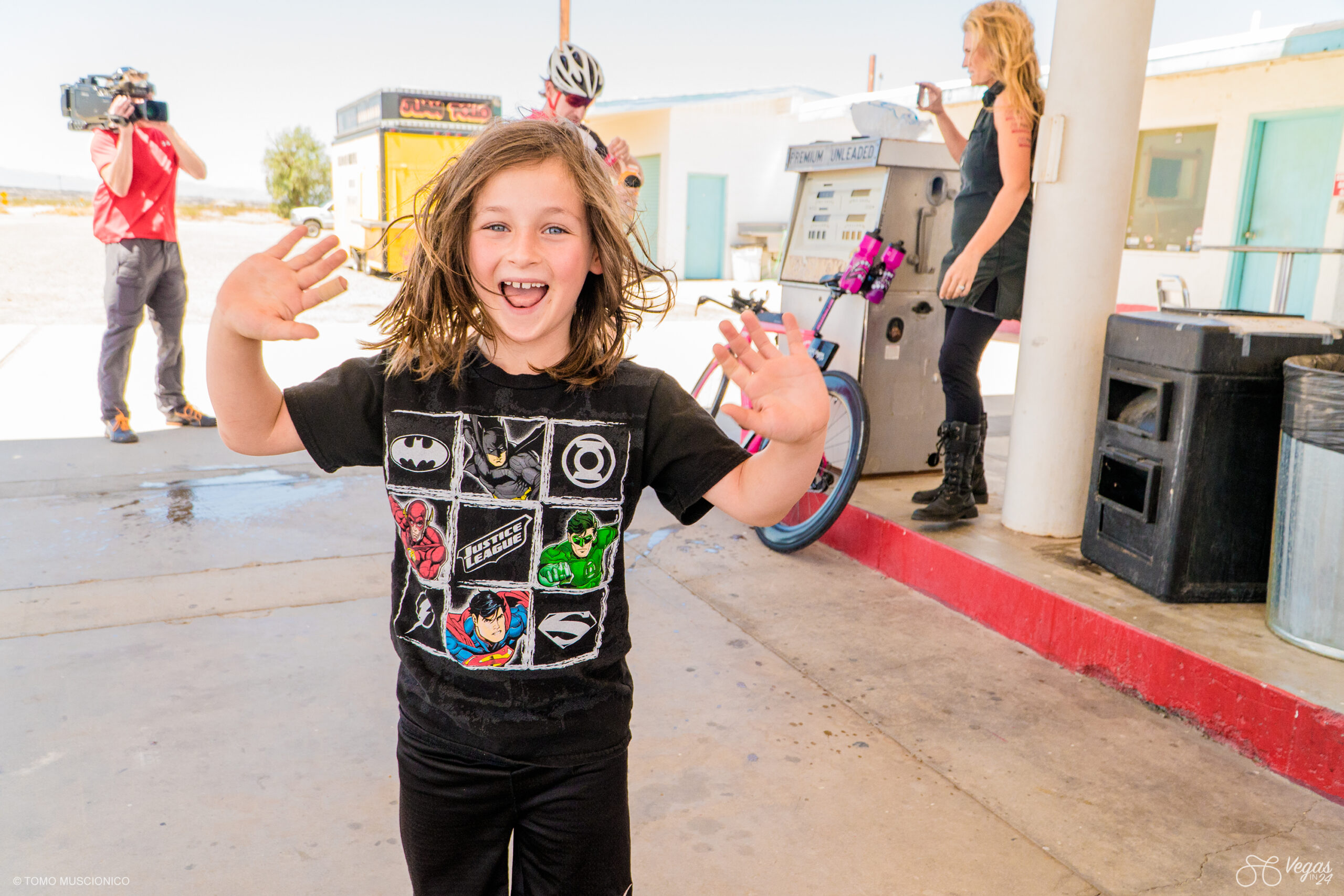 Sebastian Funk, son of Ho'Omau Endurance Founder Andy Funk, with shoulder-length brown hair, wearing a superhero t-shirt, stands outdoors Roy's Cafe in Amboy, CA during the original Vegas In 24 ultra-cycling challenge in 2016. A Garmin employee is present, documenting the 365-mile event with a large, professional camera, while Dr. Kristi Funk livestreams with her phone near a gas pump.