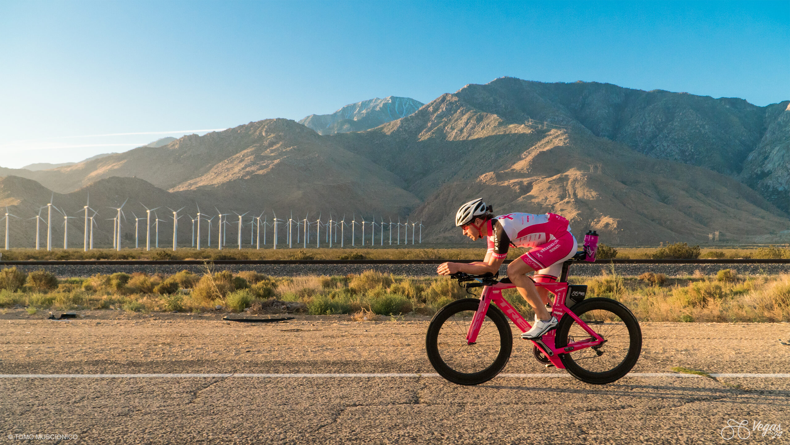 Andy Funk, who founded Ho'Omau Endurance, in a pink and white outfit rides a pink triathlon bike during the original 365-mile Vegas In 24 ultra-cycling challenge near the Palm Springs basin, with mountains and wind turbines in the background under a clear blue sky.