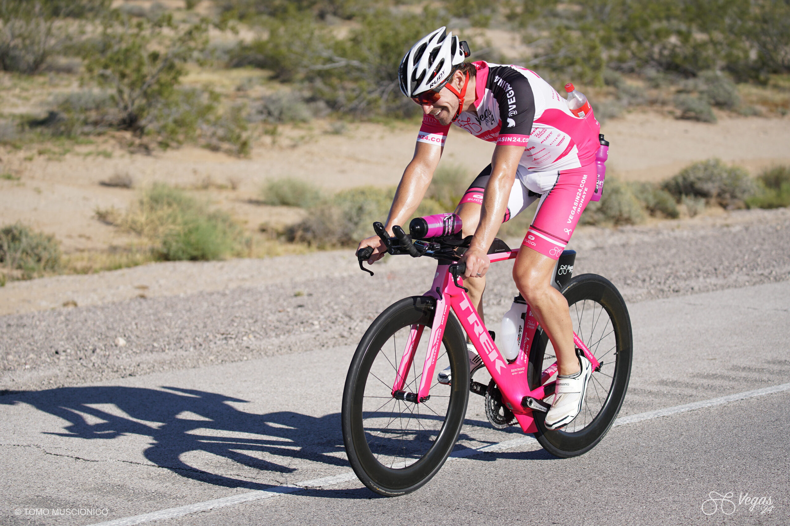 Andy Funk, CEO of Ho'Omau Endurance, in a white and pink cycling kit and helmet rides his pink Trek Speed Concept triathlon bike during the original 2016 Vegas In 24 ultra-cycling challenge. Laughing in disbelief about the never-ending long final climb to Searchlight, desert vegetation appears in the background.
