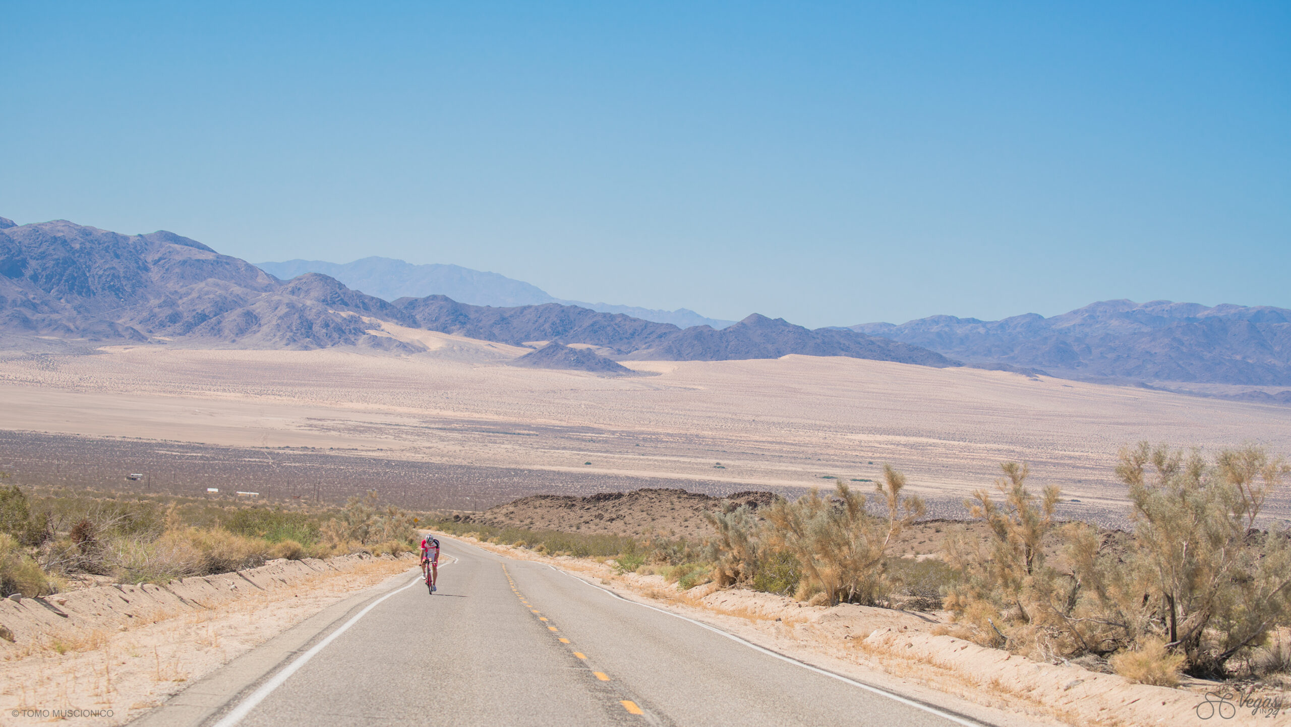 Andy Funk, Chief Ho'Omau, climbs along an empty road through a Mojave desert landscape during the original 2016 Vegas In 24 ultra-cycling challenge. Distant mountains rising beneath a clear blue sky and sparse vegetation lining the roadside.