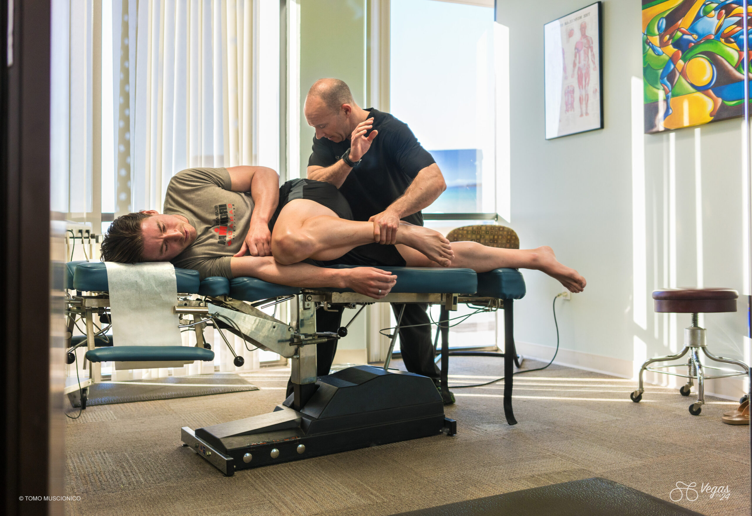 Dr. Benjamin Kleinbrodt at Brentwood Sport + Spine performs active release physical therapy work on Andy Funk, lying sideways on an exam table in a brightly lit clinic room in preparation for the the original 2016 Vegas In 24 ultra-cycling challenge, with medical charts and colorful artwork decorating the walls.