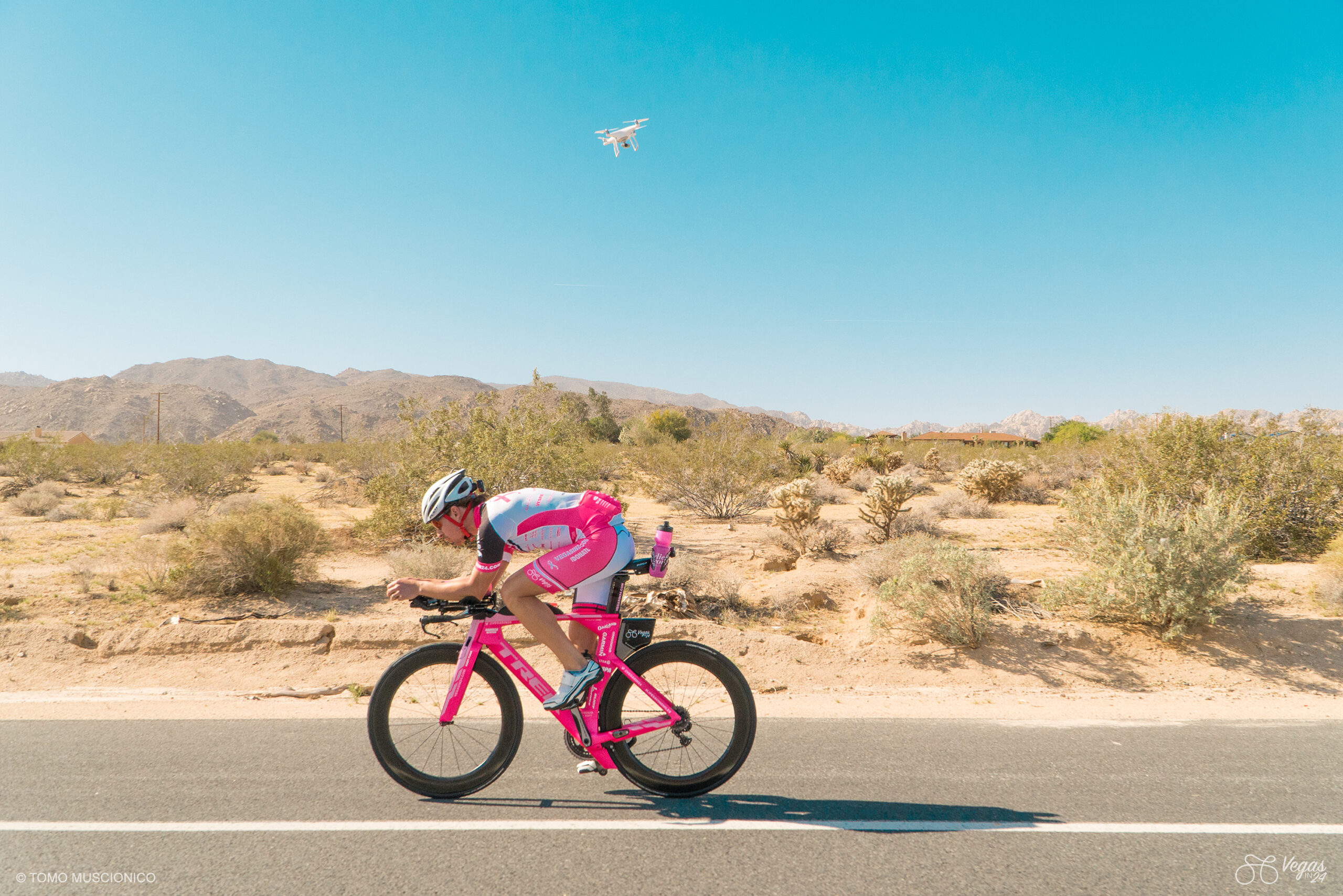 Andy Funk, Chief of Ho'Omau Endurance Co, in pink outfit and matching pink Trek Speed Concept triathlon bike along a desert road near Twenty-Nine Palms, CA, with mountains and scrub behind him. Overhead, a drone soars through the clear blue sky alongside the bike, capturing moments of the 365-mile original 2016 Vegas In 24 ultra-cycling challenge.