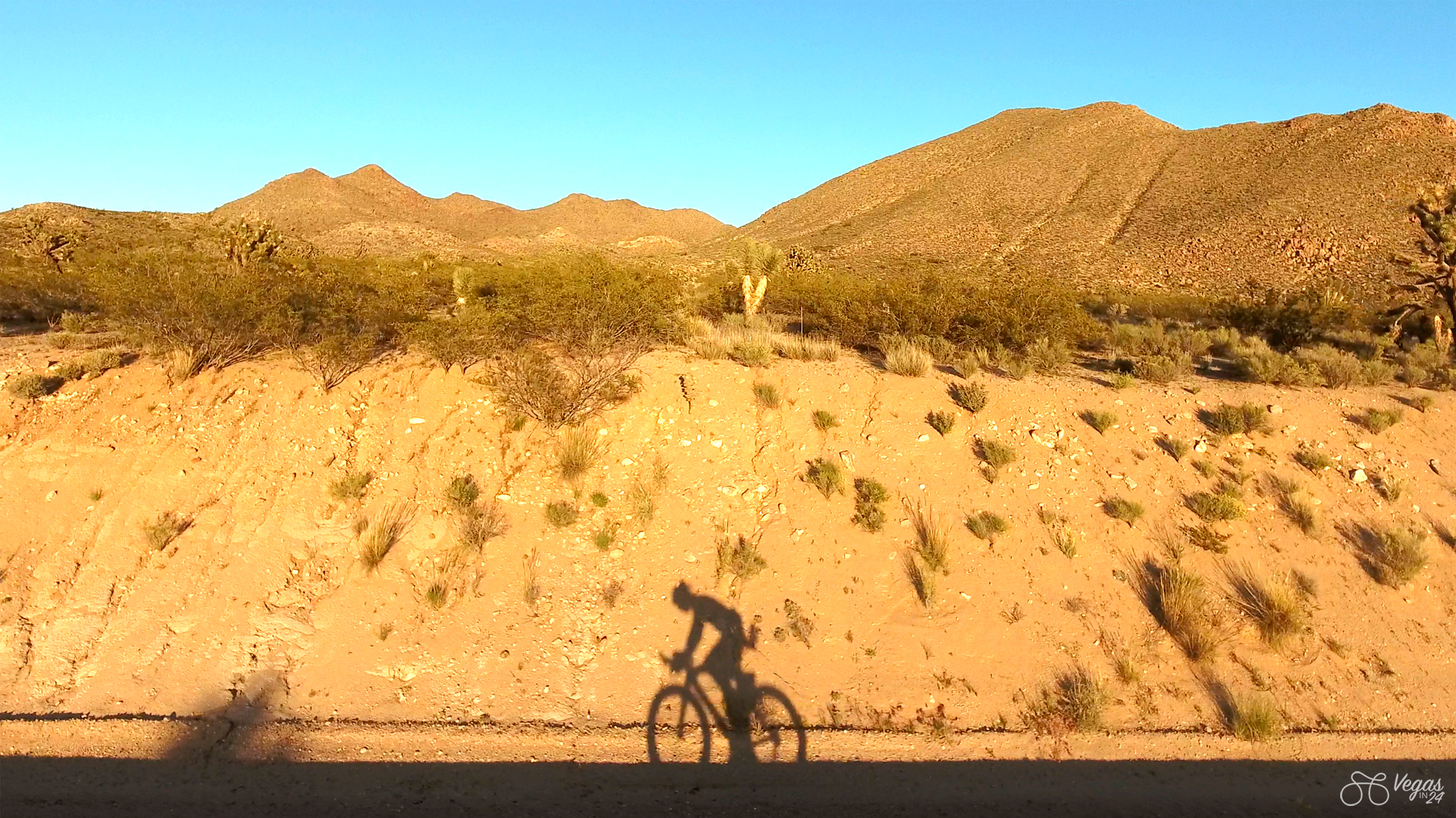 The shadow of Andy Funk, Ho'Omau Endurance chief executive, on his bicycle during the original Vegas In 24 ultra-cycling challenge in 2016, cast on a sandy embankment in a desert landscape near Searchlight, Nevada. The mountains are painted in orange from the impending sunset against and clear blue sky.