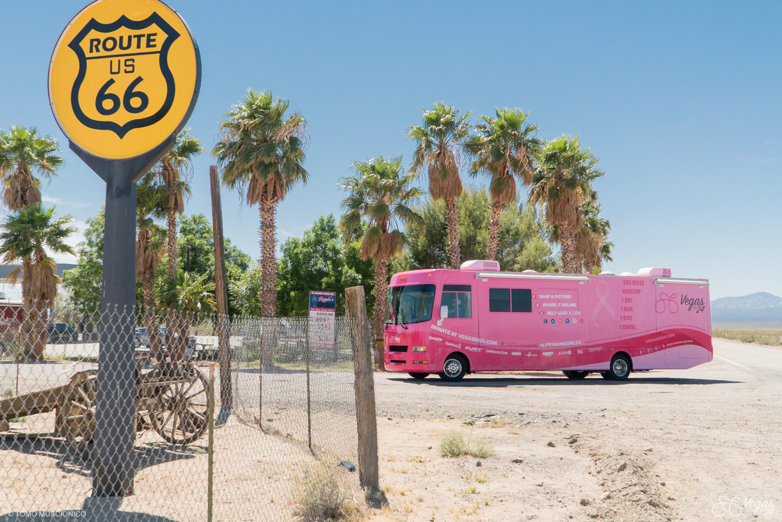 A bright pink RV entering the Fenner rest stop near palm trees and a chain-link fence with a yellow Route 66 sign in the foreground, under a clear blue sky, captured during the original Vegas In 24 ultra-cycling challenge in April 2016.