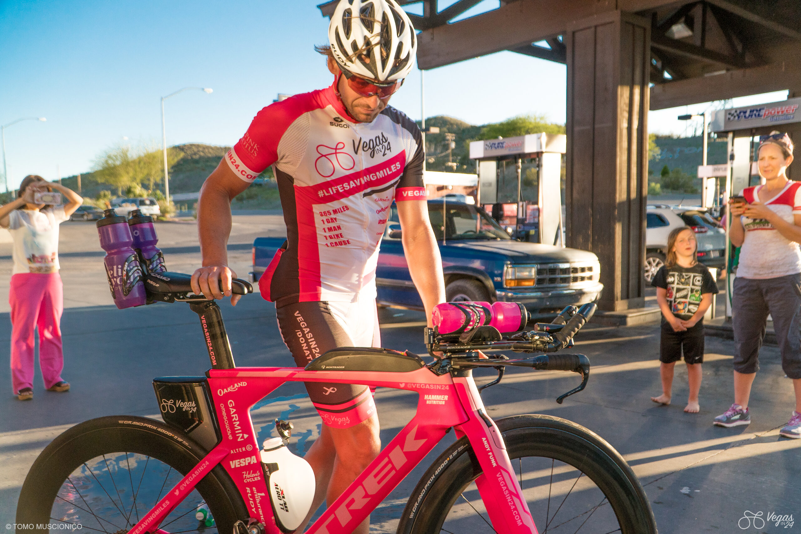 Andy Funk, Chief Executive of Ho'Omau Endurance, in a pink and white cycling kit stands beside his pink Trek bike at a gas station in Searchlight, Nevada during the 5th and final rest stop of the original Vegas In 24 ultra-cycling challenge. Dr. Helen Kang, medical director of the event, and Sebastian Funk, son of Andy Funk, watch and take photos. The sun is setting and mountains are visible in the background.