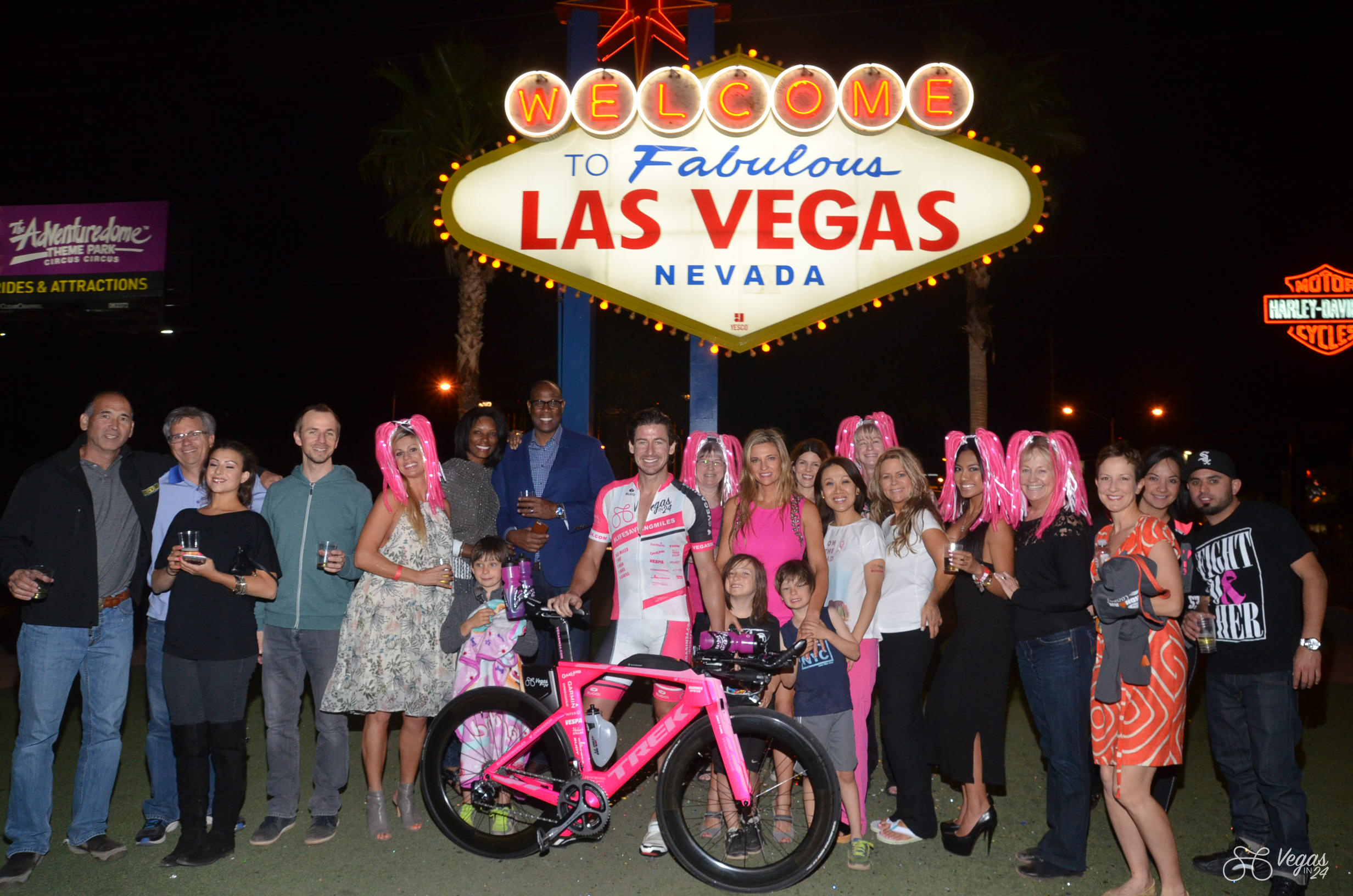 A group of friends pose smiling in front of the illuminated “Welcome to Fabulous Las Vegas Nevada” sign not long after Andy Funk completed the inaugural Vegas In 24 ultra-cycling challenge in April 2016. The group stands around Andy and his pink bicycle; many wear or hold pink items, and Dr. Kristi Funk and tje Funk triplet boys are included in the group.