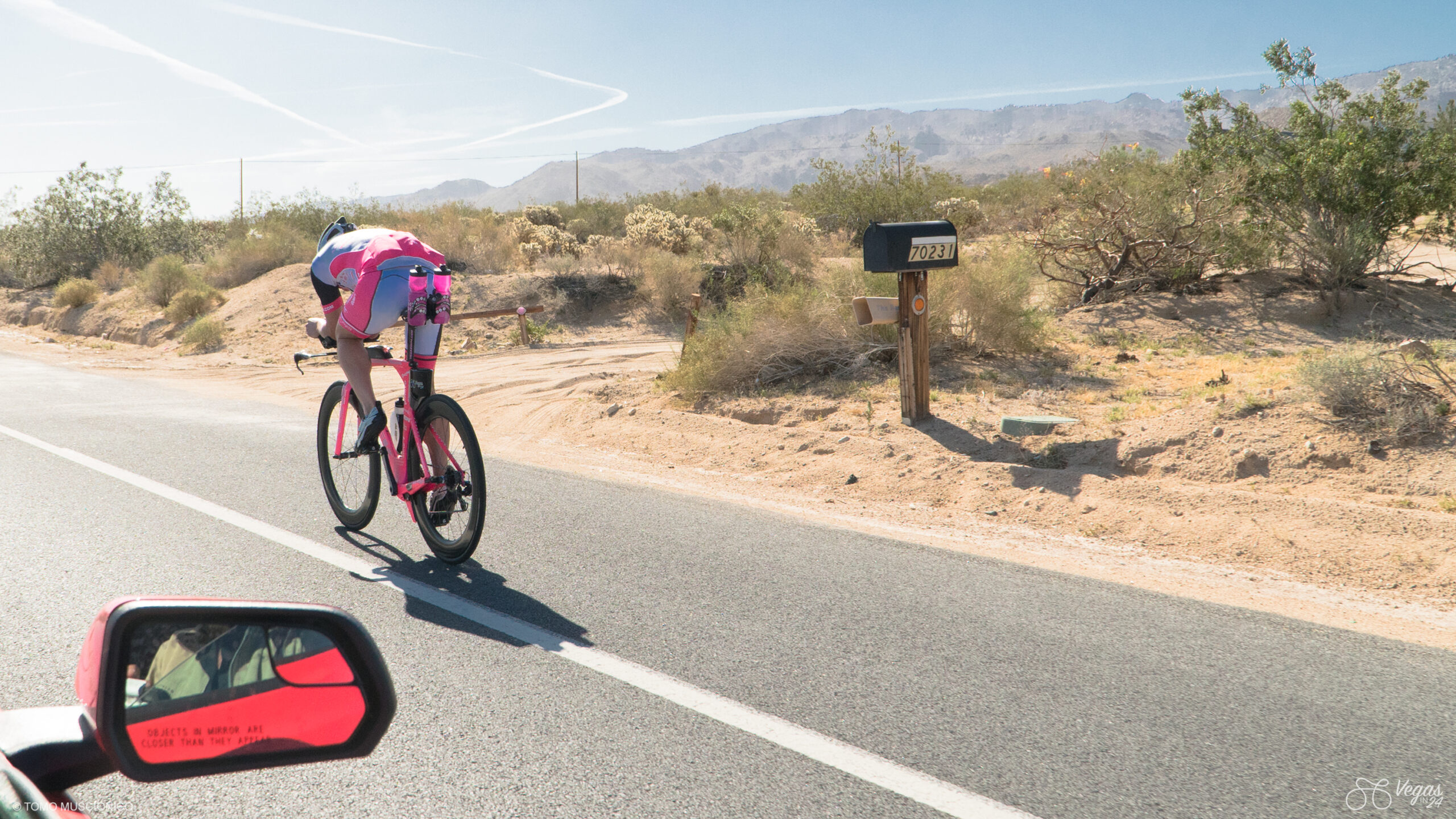 Ho'Omau Founder and CEO Andy Funk rides in his pink and white outfit on a Trek Speed Concept along a sunny desert road near Twenty-Nine Pals, California during the inaugural 2016 attempt of the Vegas In 24 ultra-cycling challenge. A mailbox and brush line the roadside, with the side mirror of the event photographers' car in the foreground and distant mountains ahead.