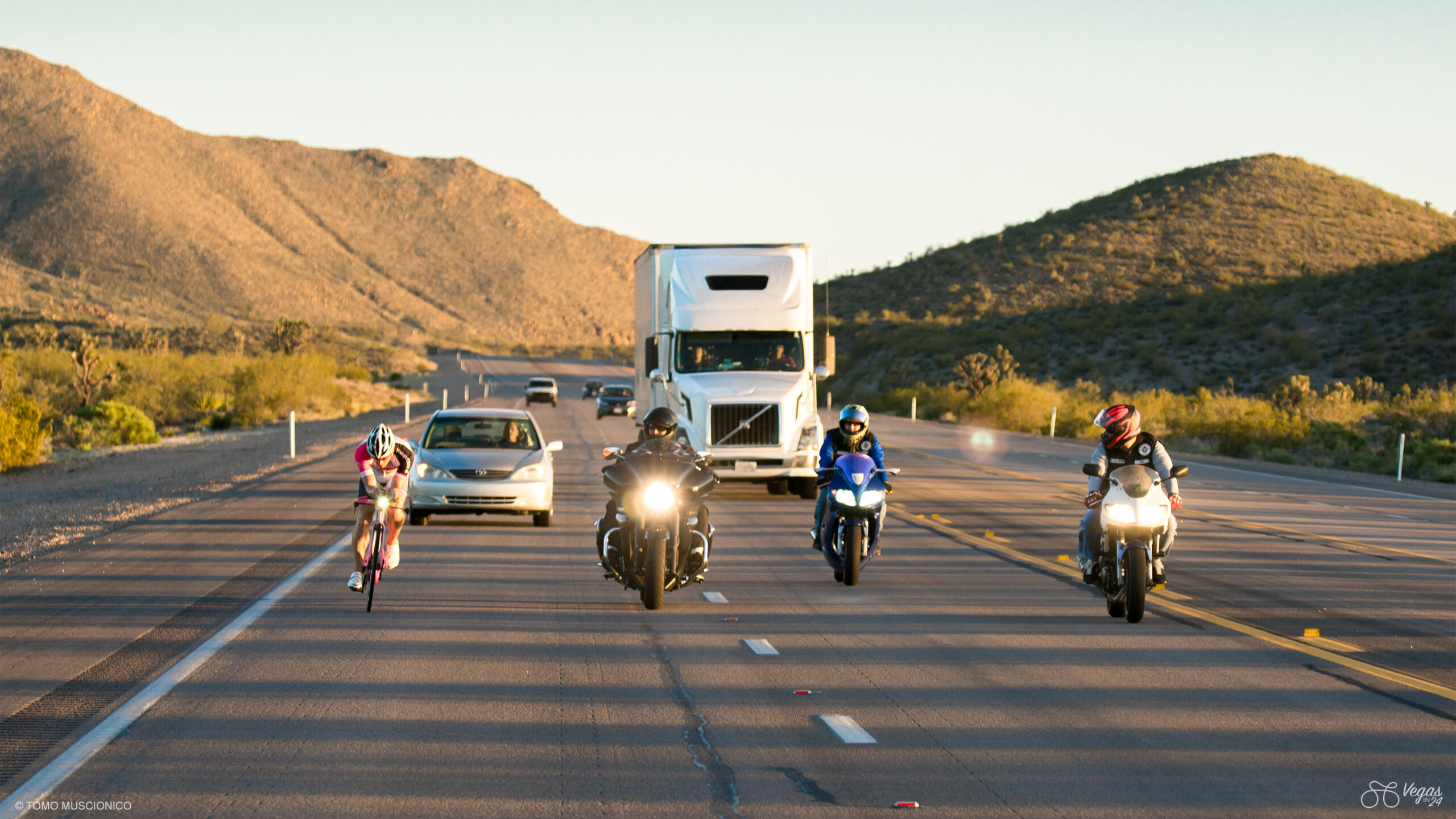 Ho'Omau Endurance founder Andy Funk embarks on the final leg, from Searchlight to Las Vegas, during the original edition of the Vegas In 24 ultra-cycling challenge in. April 2016. Riding along the left side of a highway, joined by three motorcyclists showing their respect for the effort, while a white semi-truck and cars approach in the opposite lane through the desert hills.