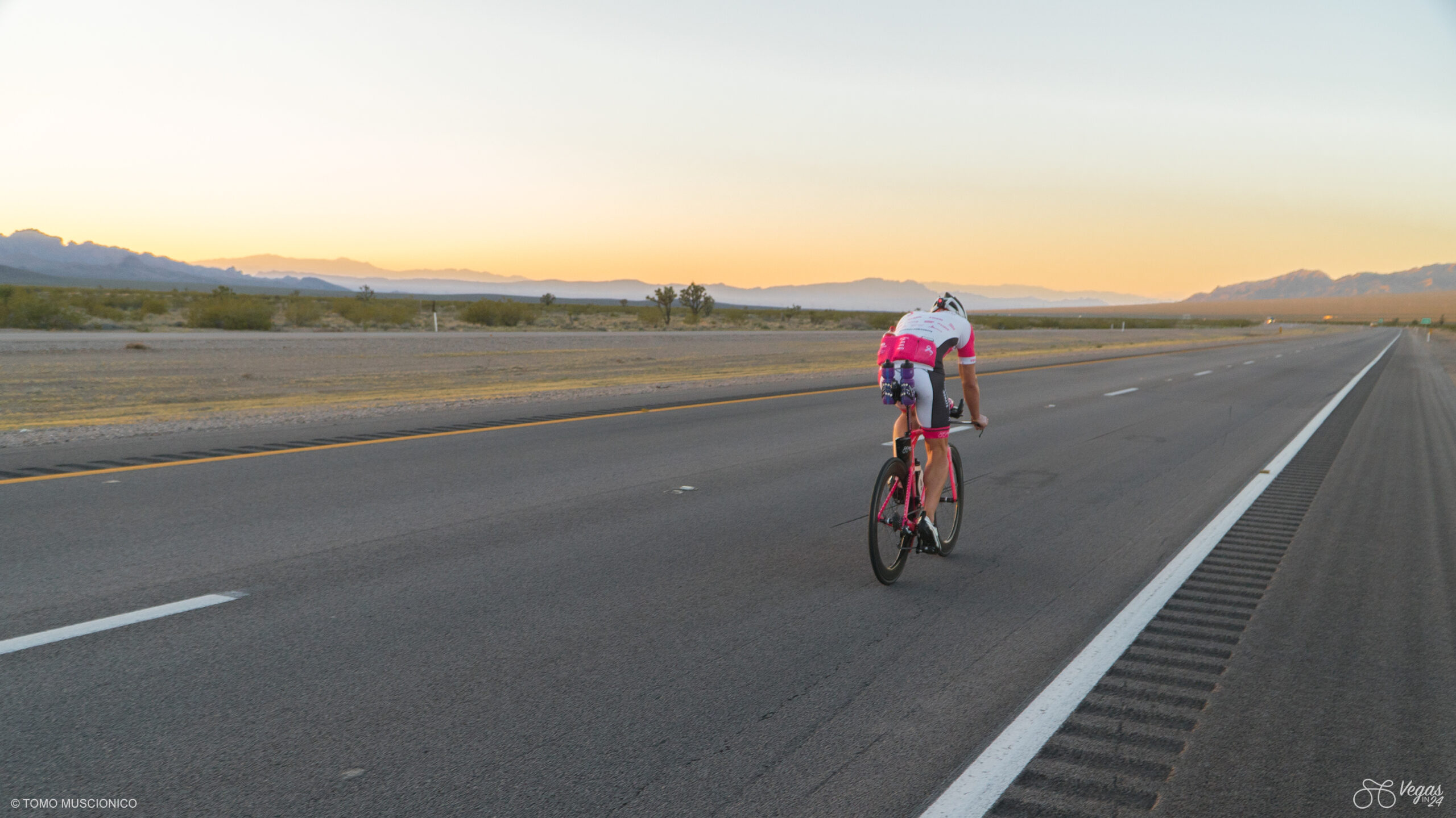 Ho'Omau Chief Andy Funk, wearing a white and pink outfit, rides his pink bike down an empty highway through a desert landscape at sunrise, embarking on the final miles towards Las Vegas during the original 365-mile Vegas In 24 ultra-cycling challenge in 2016.