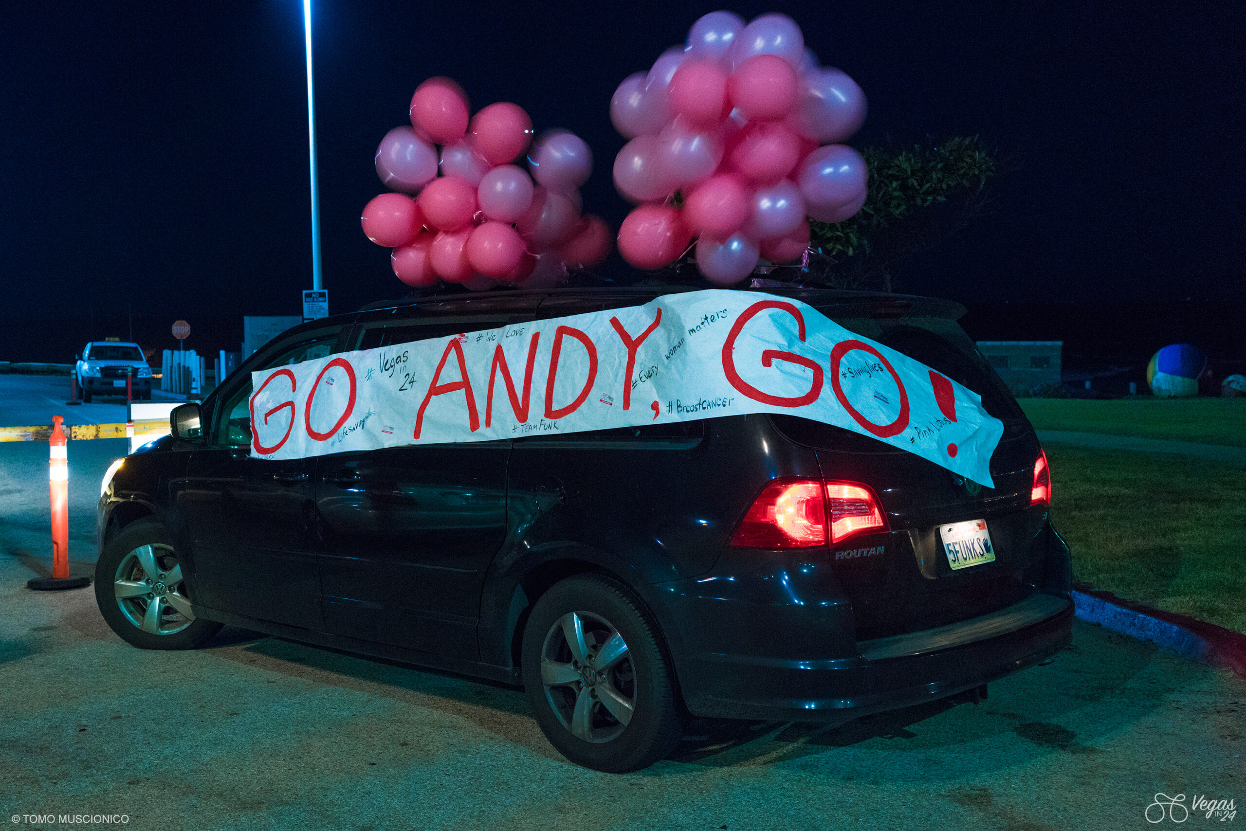 A black Minivan decorated with pink balloons and a large handmade sign reading GO ANDY GO in red letters is parked outdoors at Dockweiler Beach, California near LAX, the starting line of the original 365-mile Vegas In 24 ultra-cycling challenge. The sign is filled with handwritten messages and hashtags.