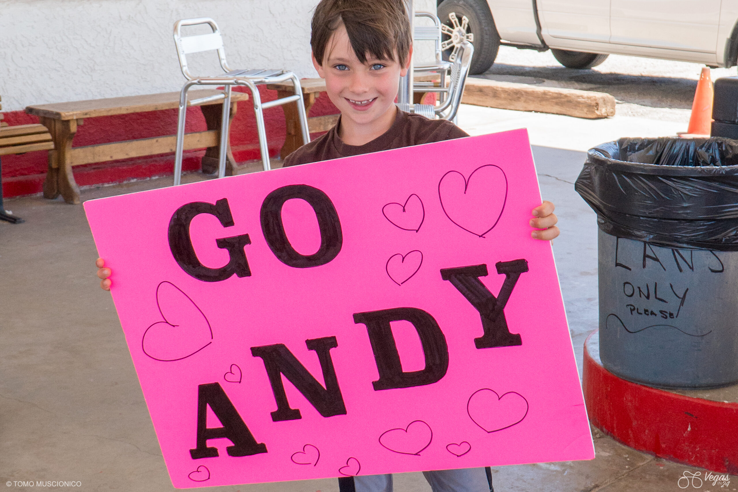 Justin Funk, son of Ho'Omau Founder Andy Funk, holds a large bright pink sign that reads GO ANDY in bold black letters, decorated with hand-drawn hearts—cheering on Andy during the inaugural 2016 Vegas In 24 ultra-cycling challenge. Justin stands outdoors at a gas station near a trash can and some metal chairs.