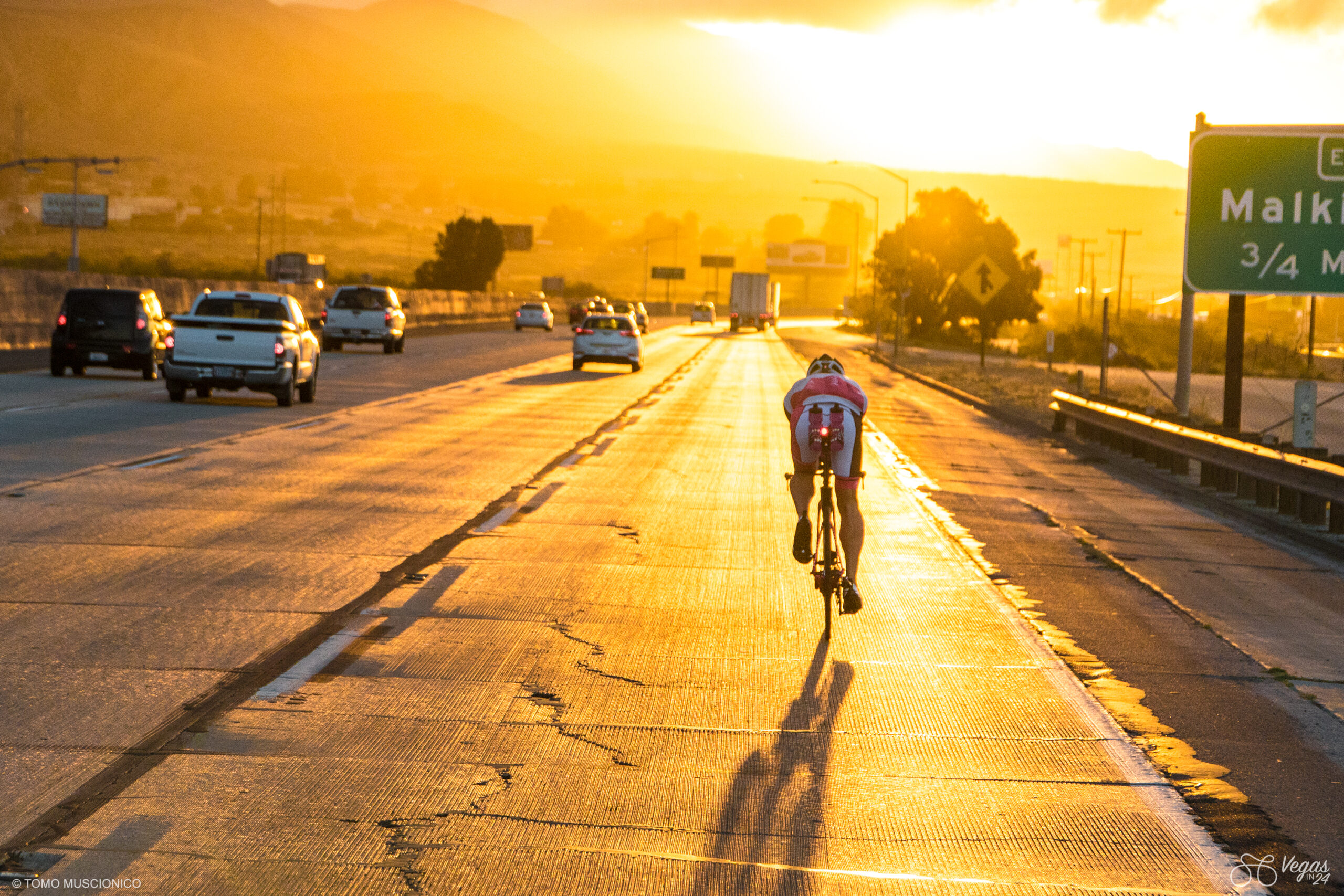 Ho'Omau Endurance Chief Andy Funk rides along a sunlit I-10 at sunrise during the first-ever Vegas In 24 ultra-cycling challenge attempt in April 2016, casting a long shadow on the road. Cars pass in the opposite lane, and a green road sign stands visible on the right side.