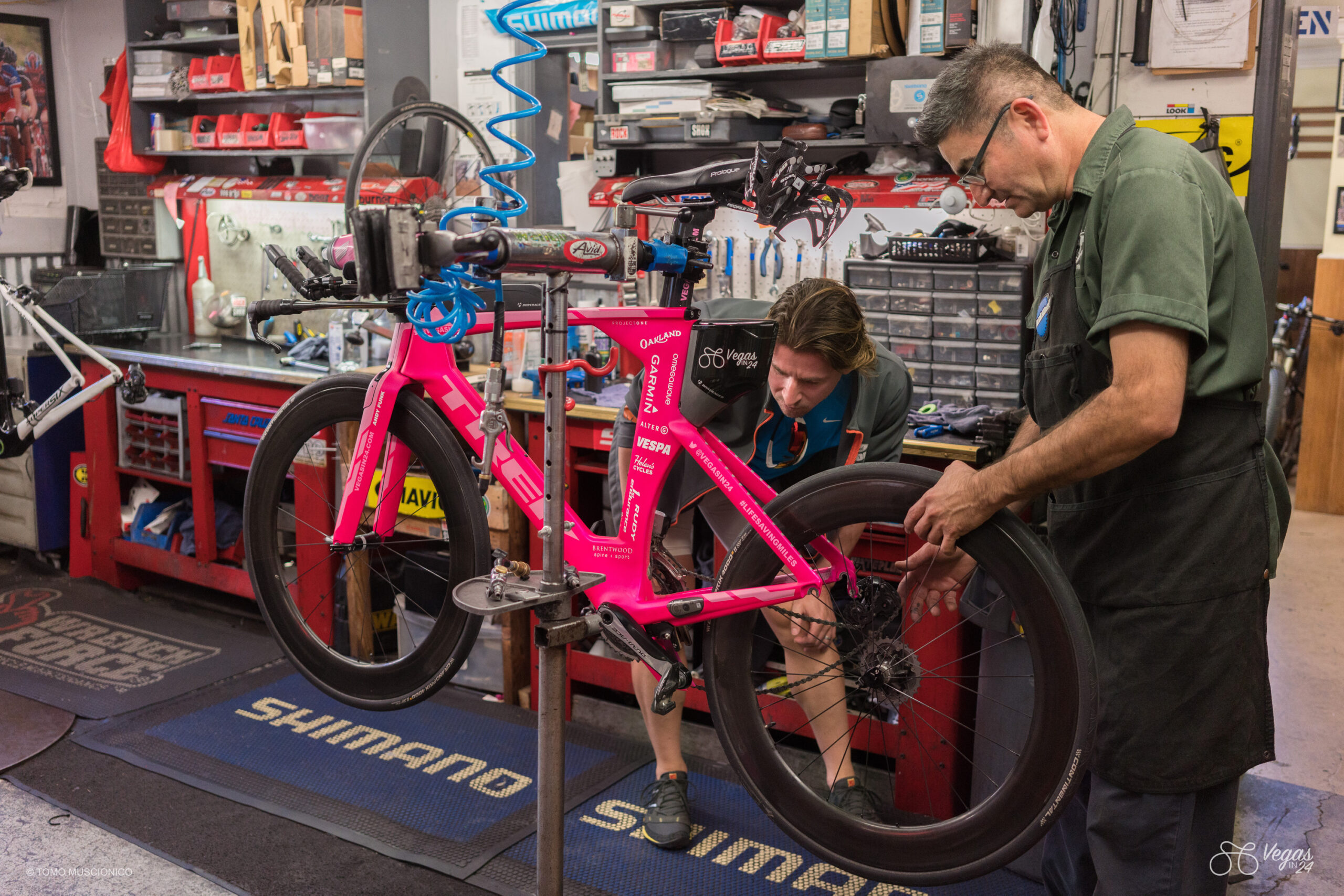 Andy Funk, Founder & Chief of Ho'Omau Endurance, observes a bike mechanic during the final bike check and tune-up prior to attempting the first-ever Vegas In 24 365-mile cycling challenge. Situated in a well-equipped workshop and surrounded by tools and gear, the mechanic adjusts the back wheel while Andy checks the bike.