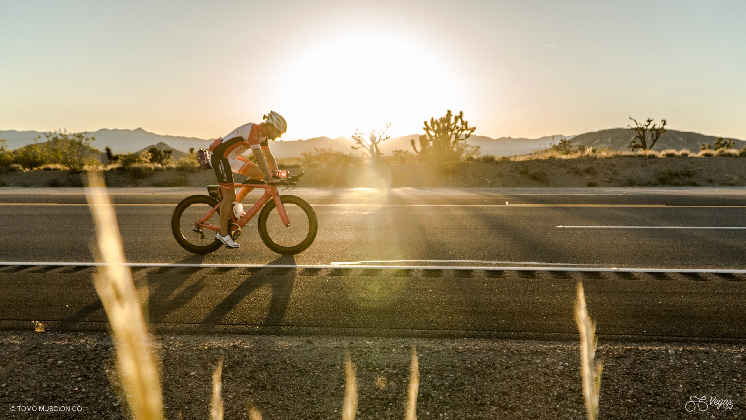 Ho'Omau founder and athlete Andy Funk rides ride along an empty Highway 95 at sunset, mountains and desert vegetation in the background, during the final hours of the first-ever 365-mile Vegas In 24 ultra-cycling challenge. Sunlight casts a warm glow and lens flare across the scenic landscape.